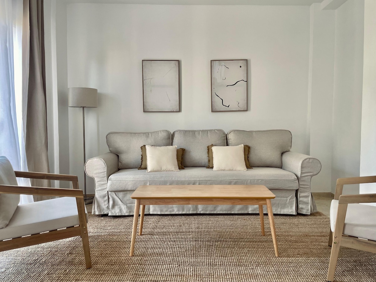 The living area features a light-colored sofa with decorative pillows, positioned between two minimalist armchairs. A wooden coffee table sits in front, and two abstract framed artworks adorn the wall. Natural light filters in through adjacent windows, highlighting the soft textures of the space.