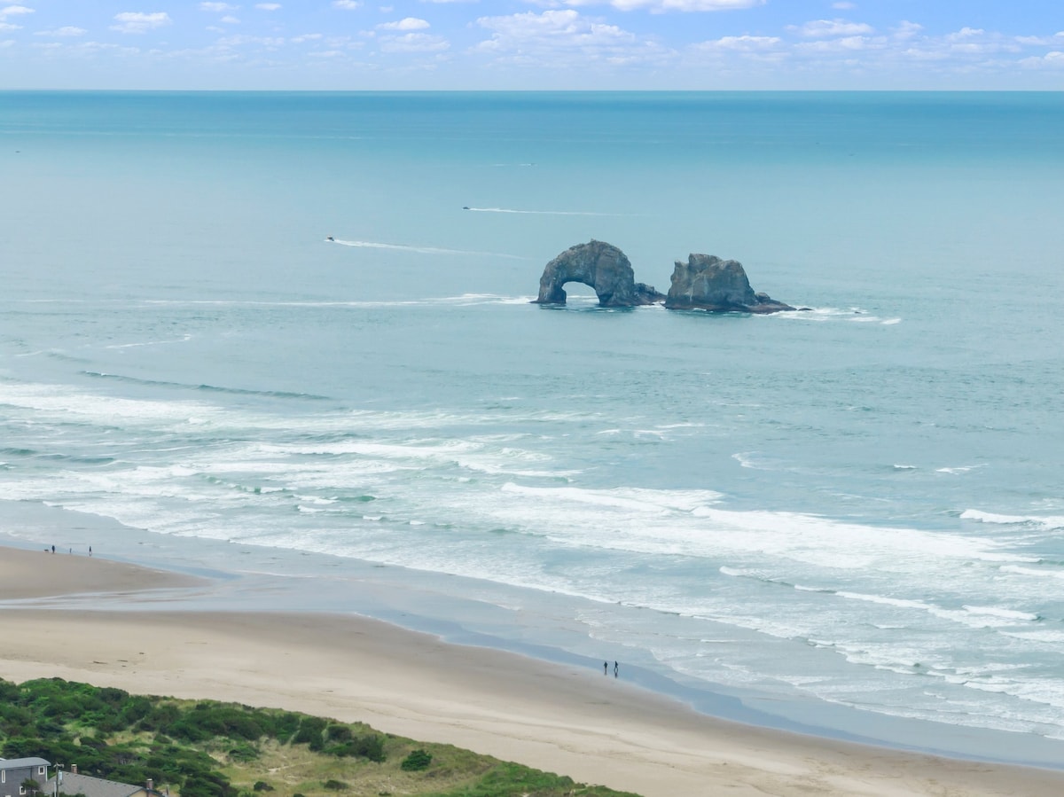 The image captures a serene coastal view where the ocean meets the sandy beach. Two distinct rock formations can be seen jutting from the water, framed by gentle waves and a clear sky filled with soft clouds.