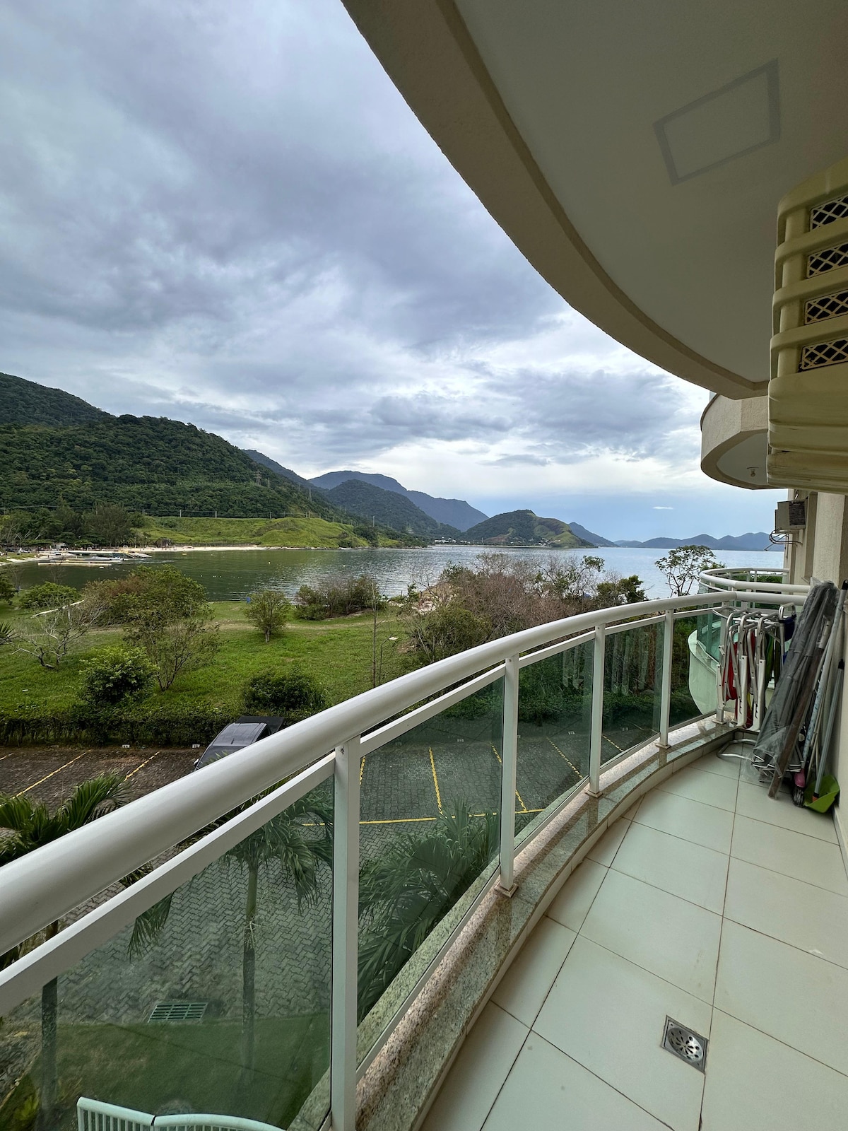 A curved balcony is showcased, offering an unobstructed view of lush green hills and a serene body of water. The railing provides safety while allowing natural light and fresh air into the space. Surrounding areas feature grass and palm trees, enhancing the natural setting.