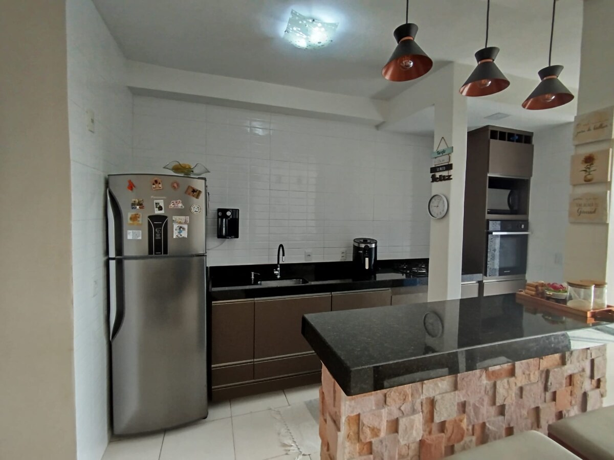 A well-equipped kitchen features modern appliances including a stainless-steel refrigerator, built-in oven, and stovetop. Dark cabinets are complemented by a sleek black countertop. Pendant lights hang above a breakfast bar with stone detailing, providing a welcoming space for casual dining.