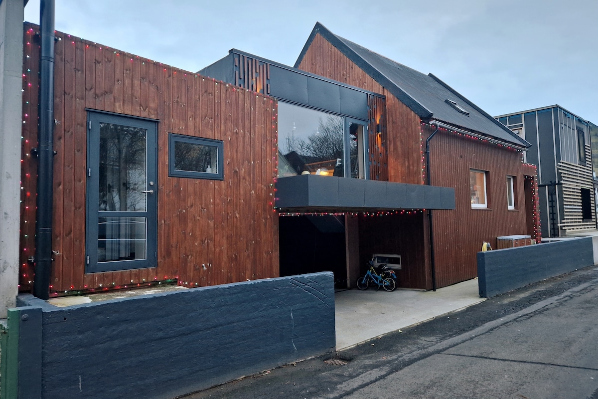 A renovated building featuring a wooden exterior with modern elements. Large windows allow natural light to enter, while decorative lights enhance the entrance. The structure combines traditional and contemporary styles, with a spacious area below an overhang used for outdoor seating or storage.