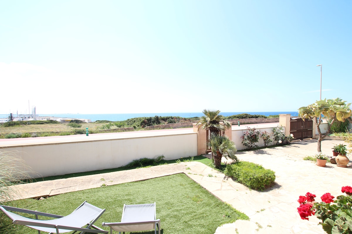 A spacious outdoor area features a well-maintained garden with laid stones and patches of grass. Two loungers are positioned for relaxation, while flowering plants and palm trees add greenery. A view of the sea can be seen in the background, enhancing the natural setting.