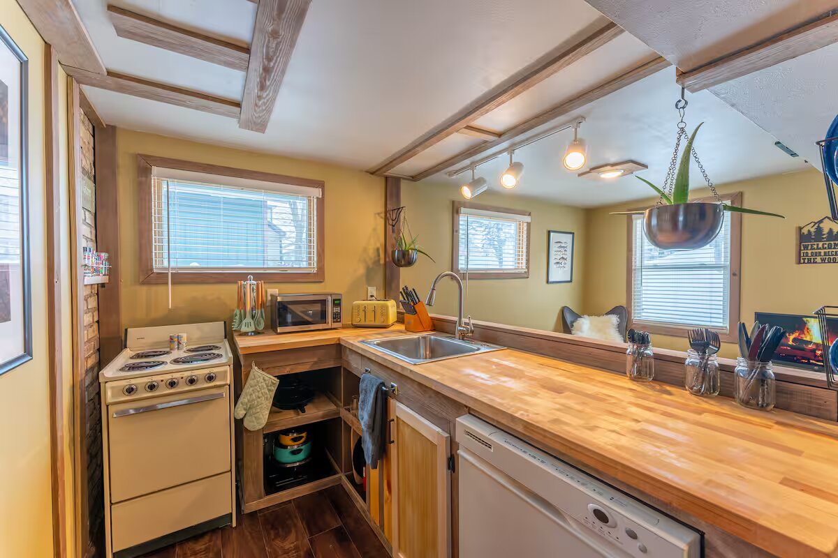 The kitchen features an inviting layout with wooden countertops and modern appliances. Natural light enters through the windows, illuminating the space. An assortment of kitchenware is neatly arranged, and a cozy seating area is visible in the background, highlighting the cabin's warmth.