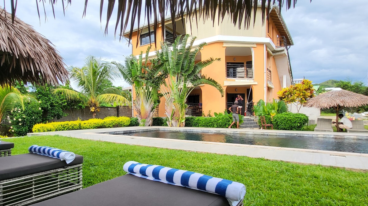 A modern two-story house is set amongst lush greenery, with palm trees and well-maintained landscaping surrounding a tranquil pool. Sun loungers with blue and white towels offer a relaxing area by the water, ideal for enjoying the peaceful outdoor space.