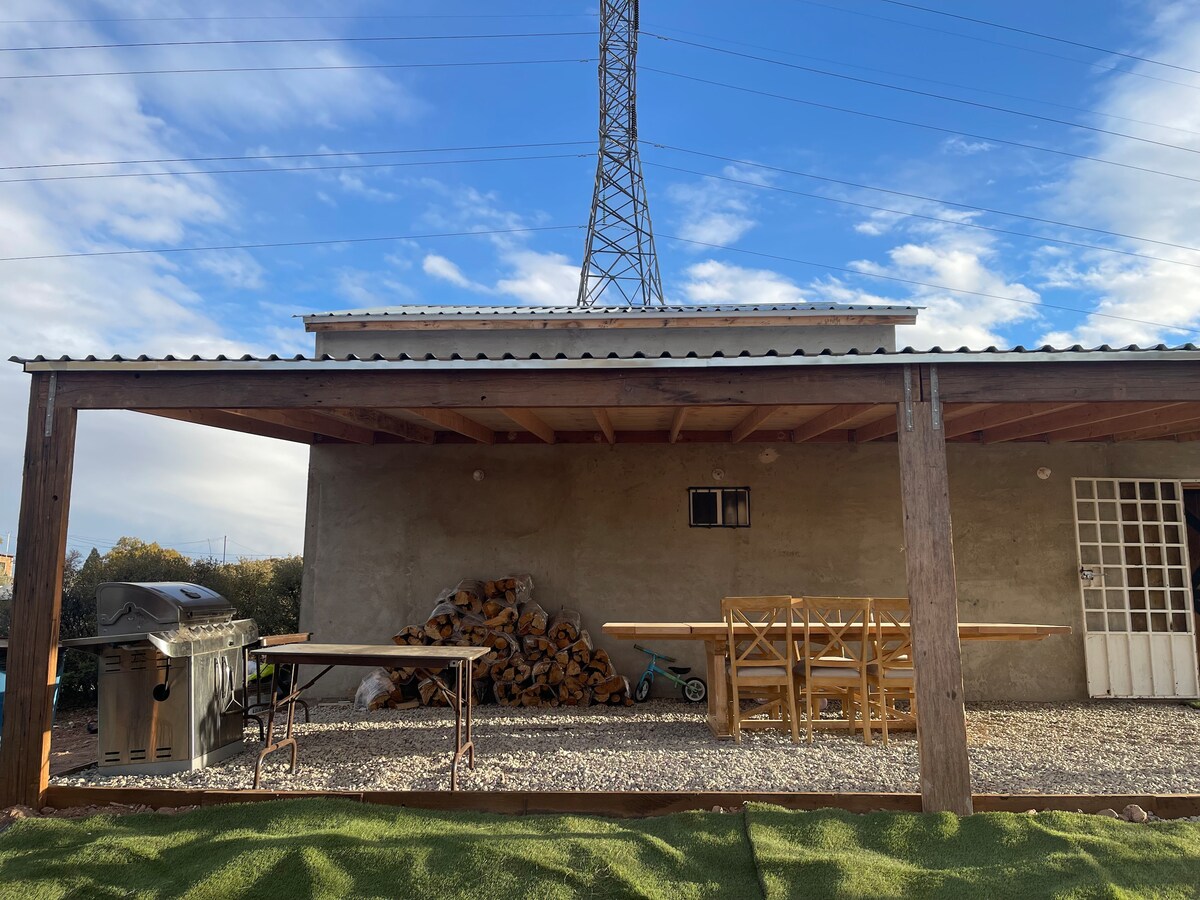 An outdoor patio area is visible, featuring a barbecue grill and wooden tables. Stacked firewood is placed nearby, and the ground is covered with gravel. A large building is seen in the background, topped with a metal roof against a blue sky with some clouds.