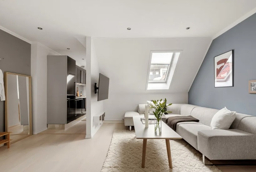 A spacious living area features a light-colored sectional sofa and a coffee table placed on a soft rug. A wall-mounted television is positioned across from the seating. Natural light enters through a skylight, illuminating the room's neutral tones. An adjacent kitchen area can be seen.
