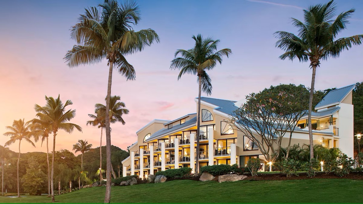 The resort building is framed by palm trees, showcasing a modern architectural design. Balconies with glass railings are visible on the upper levels. Soft hues of sunset illuminate the sky, creating a serene ambiance. Lush greenery surrounds the property, contributing to the tropical setting.