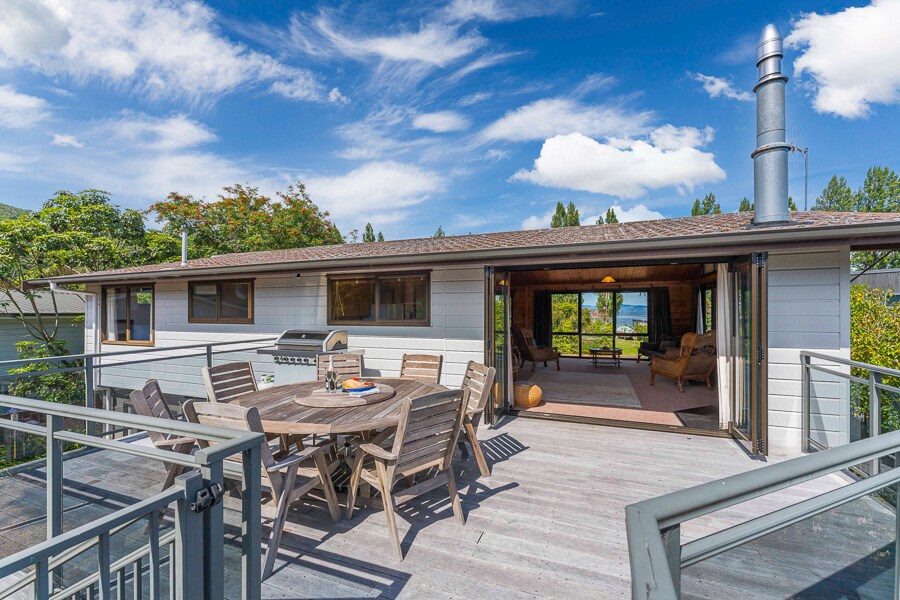 A spacious outdoor deck features a dining table with seating for six and a barbecue grill, overlooking a lush yard. Large glass doors lead into a bright living area, revealing views of the surrounding trees and lake. A chimney pipe extends from the roof, contributing to the home's rustic charm.