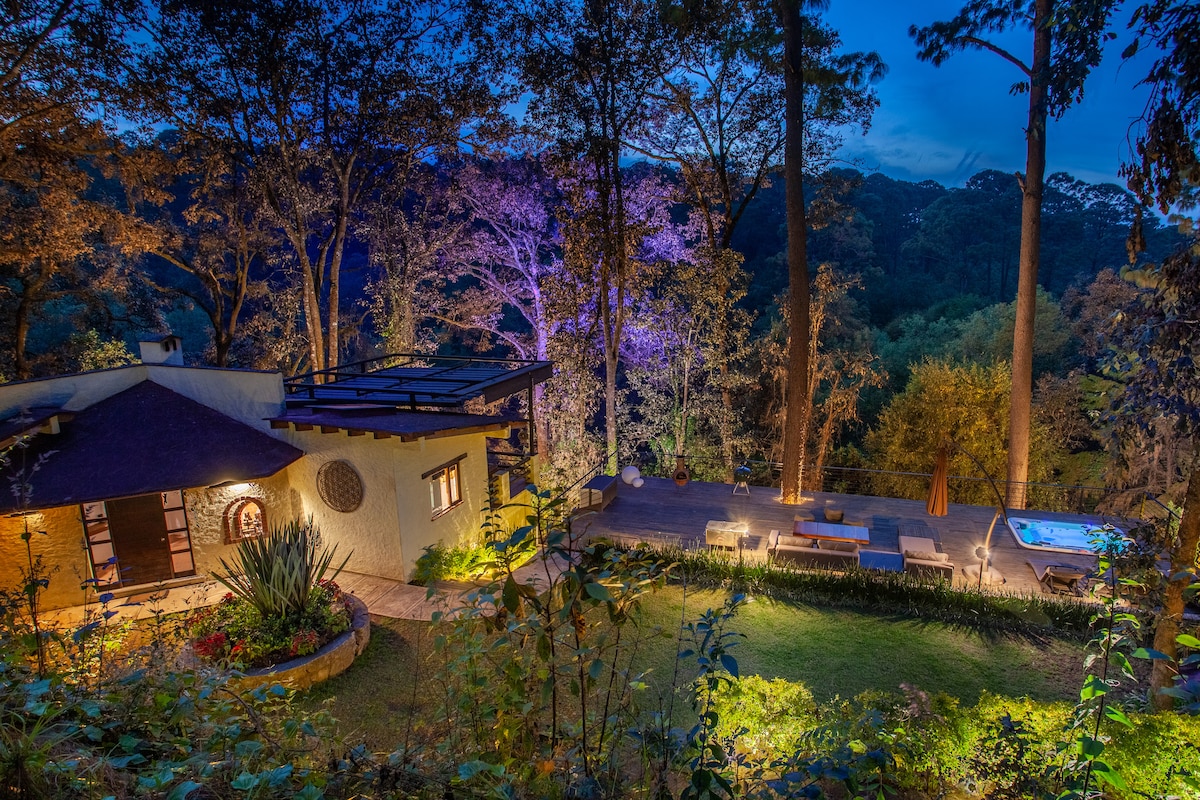 An evening view of the property showcases a spacious outdoor area illuminated by ambient lighting, featuring a jacuzzi and sun loungers. The distinct architecture of the house is complemented by surrounding trees, creating a serene atmosphere in the natural setting.
