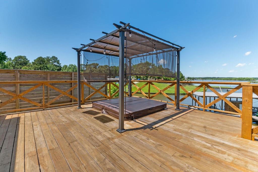 An elevated deck features a spacious lounging area shaded by a canopy. Wooden planks create a warm ambiance, with glimpses of the surrounding green landscape and water visible beyond the railing.