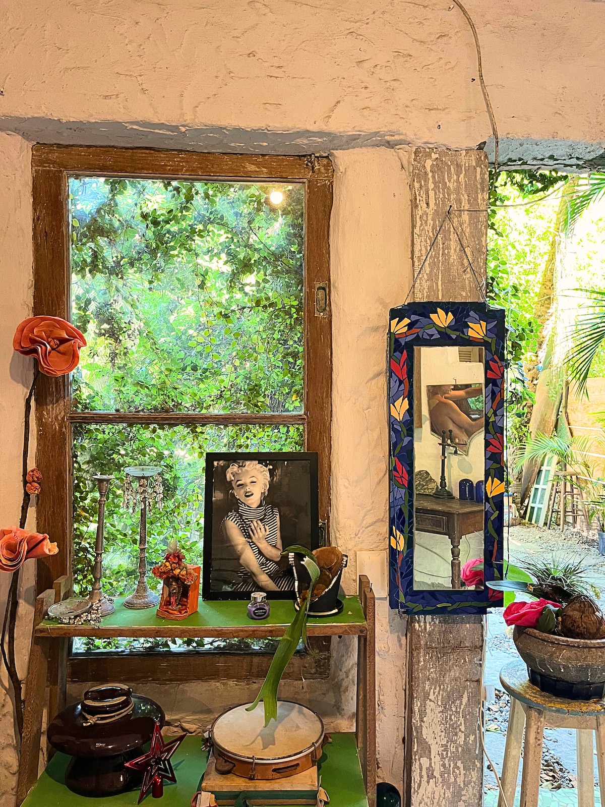A rustic interior corner is shown, featuring a wooden window framed by green foliage outside. Decorative items, including a mirror, art pieces, and vases, create an eclectic display on a green shelf, reflecting an artistic atmosphere within the home.