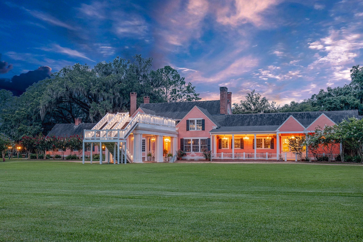 The exterior of a historical building is presented, showcasing a peach-colored facade with white trim. Large windows reflect the evening light, while a spacious porch adds to the welcoming feel. Surrounding grassy areas are complemented by mature trees and evening sky colors.