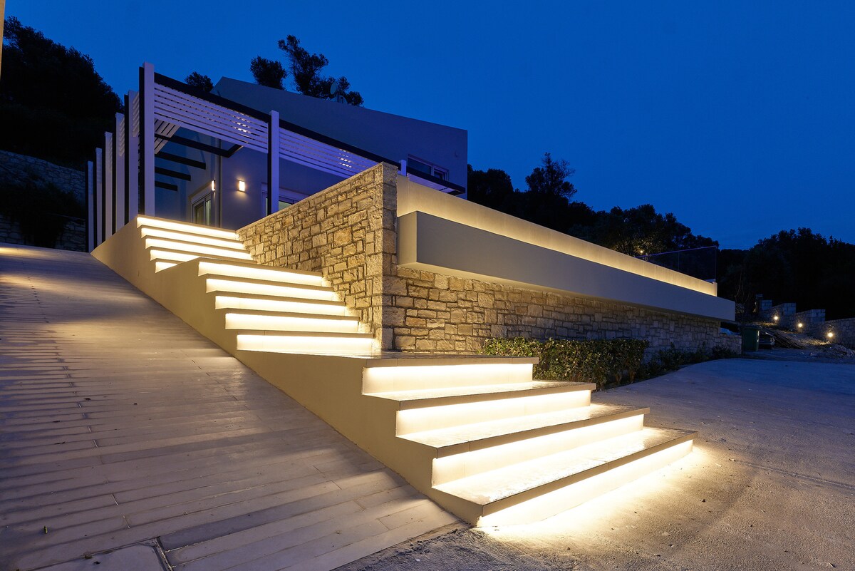 A softly lit entrance features illuminated steps leading to the villa, with a stone wall adding texture to the inviting pathway. Surrounding greenery is visible in the background, enhancing the serene ambiance of the evening view.