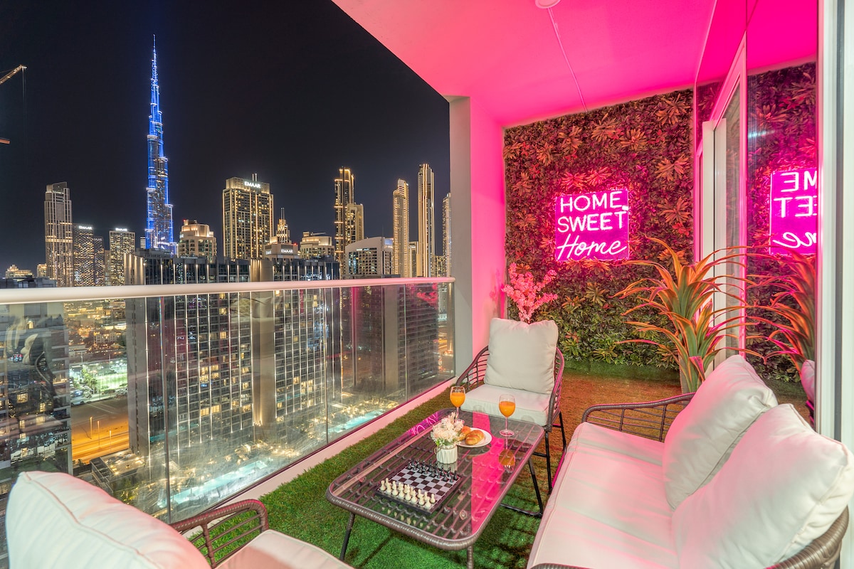 A balcony setting is presented with comfortable seating and a vibrant city skyline view, featuring the Burj Khalifa illuminated at night. Decorative greenery and a neon sign stating 'HOME, SWEET HOME' add a modern touch, creating an inviting space for relaxation.