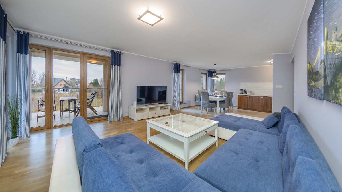A spacious living area features a large, comfortable sectional sofa in blue, paired with a glass coffee table. Natural light floods through wide glass doors that open to a balcony, with dining chairs and a table visible in the distance.