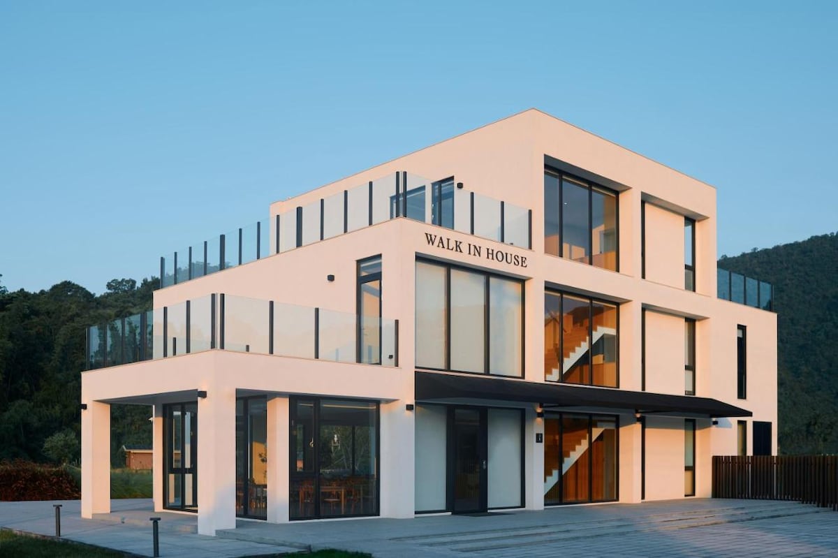 A modern three-story building is shown, featuring sleek lines and large glass windows. The structure's design includes multiple outdoor balconies with glass railings, providing open views of the surrounding landscape. The entrance is adorned with a welcoming sign, adding to the inviting aesthetic.