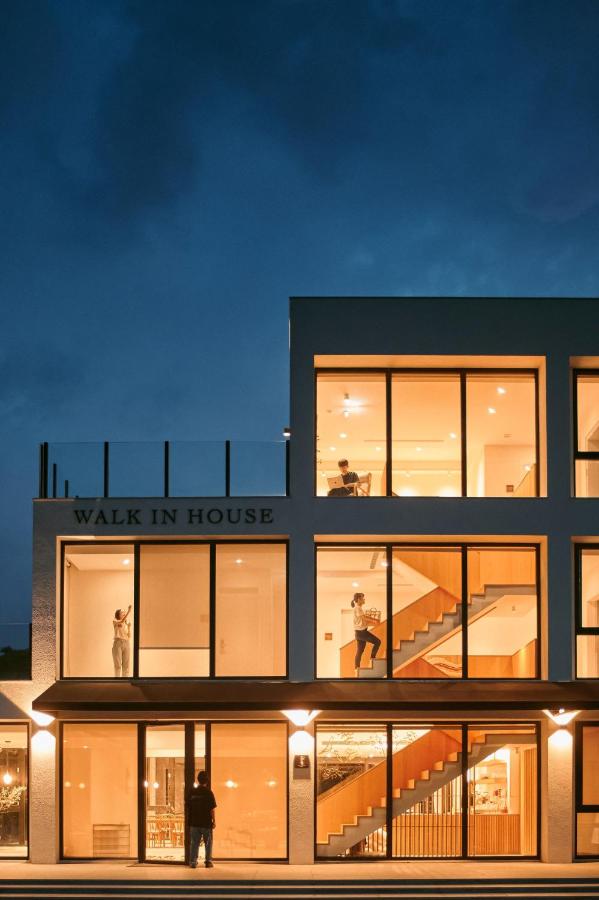 The exterior of the Walk In House is presented at twilight, illuminated by warm lights. Large glass windows reveal the interior, showcasing a staircase and guests engaged in activities. The modern architecture emphasizes clean lines and a welcoming design.