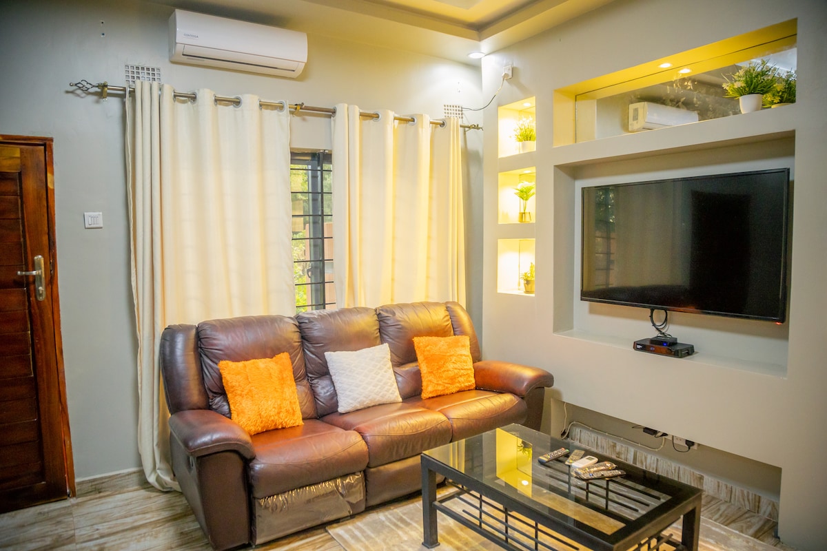 A cozy lounge area features a dark leather sofa adorned with orange and white cushions. A glass coffee table sits in front of the sofa, and light-colored curtains frame the window. A wall-mounted television is displayed above a sleek media unit with decorative plants.