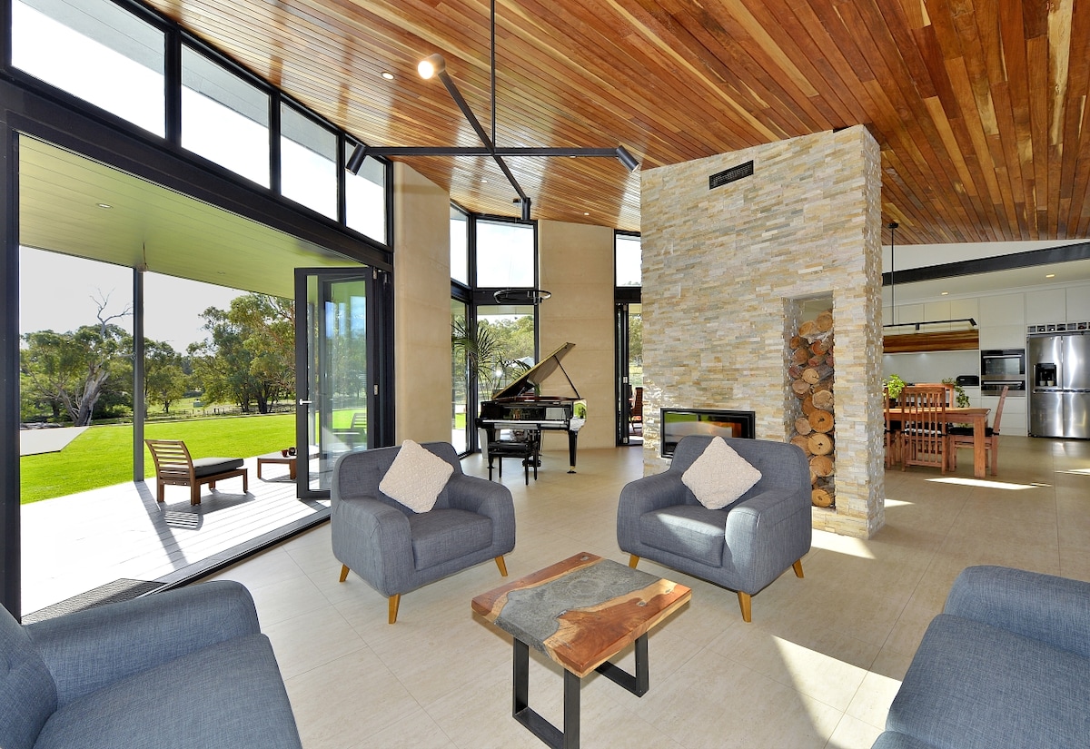 An open living area features two gray sofas with decorative pillows and a wooden coffee table. Large glass doors provide views of the lush outdoor landscape. A grand piano is situated near a stone fireplace, enhancing the connection between indoor and outdoor spaces.