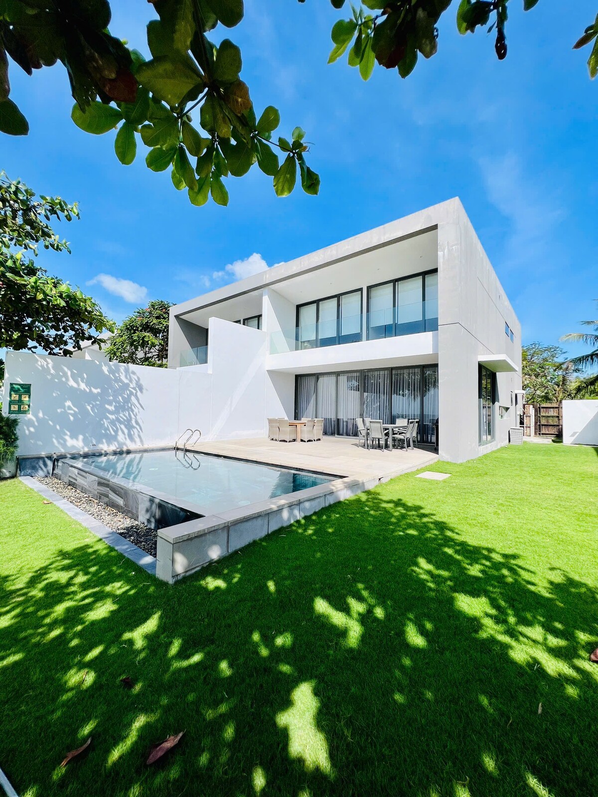 The modern villa features a clean, minimalist design with large glass windows that reflect the surrounding greenery. A private swimming pool sits adjacent to the grassy area, while shaded outdoor seating allows for relaxation. The bright blue sky enhances the serene atmosphere of the property.