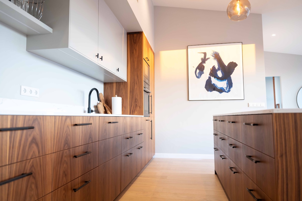 A modern kitchen space features elegant wood cabinetry with ample storage and sleek black hardware. A striking piece of wall art adds a touch of character. Soft lighting enhances the bright ambience, while a minimalist design promotes an uncluttered atmosphere.
