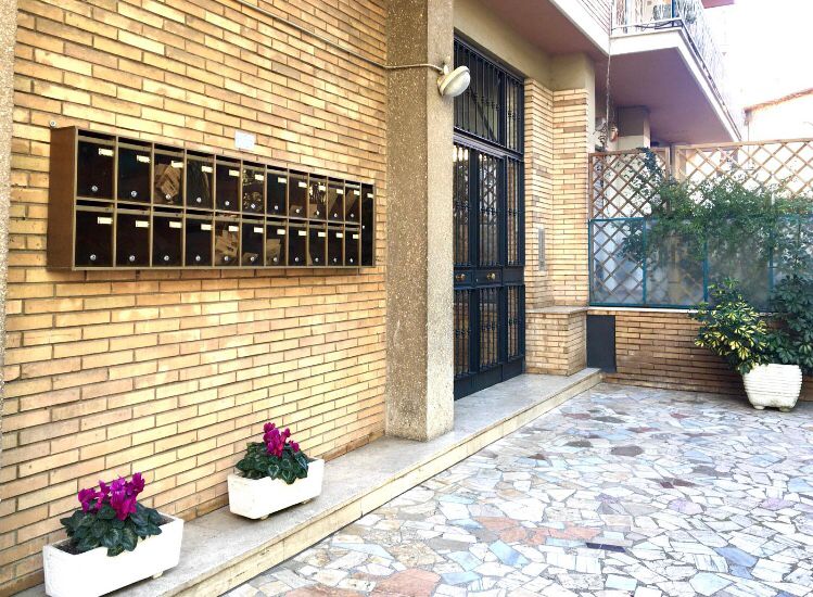 The entrance of the condominium is shown, featuring a brick facade. A row of mailboxes is mounted on the wall, beside a glass door. The pathway is decorated with stone tiles and is lined with planters containing vibrant purple flowers.