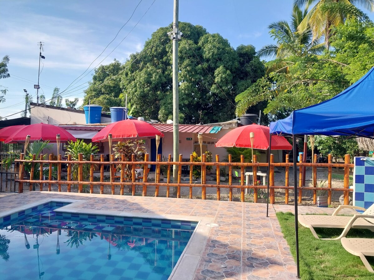A refreshing swimming pool is surrounded by a stone patio, featuring lounge chairs and bright red umbrellas for shade. The backyard includes green plants and trees, adding natural decor, while simple structures with colorful roofs are visible in the background.