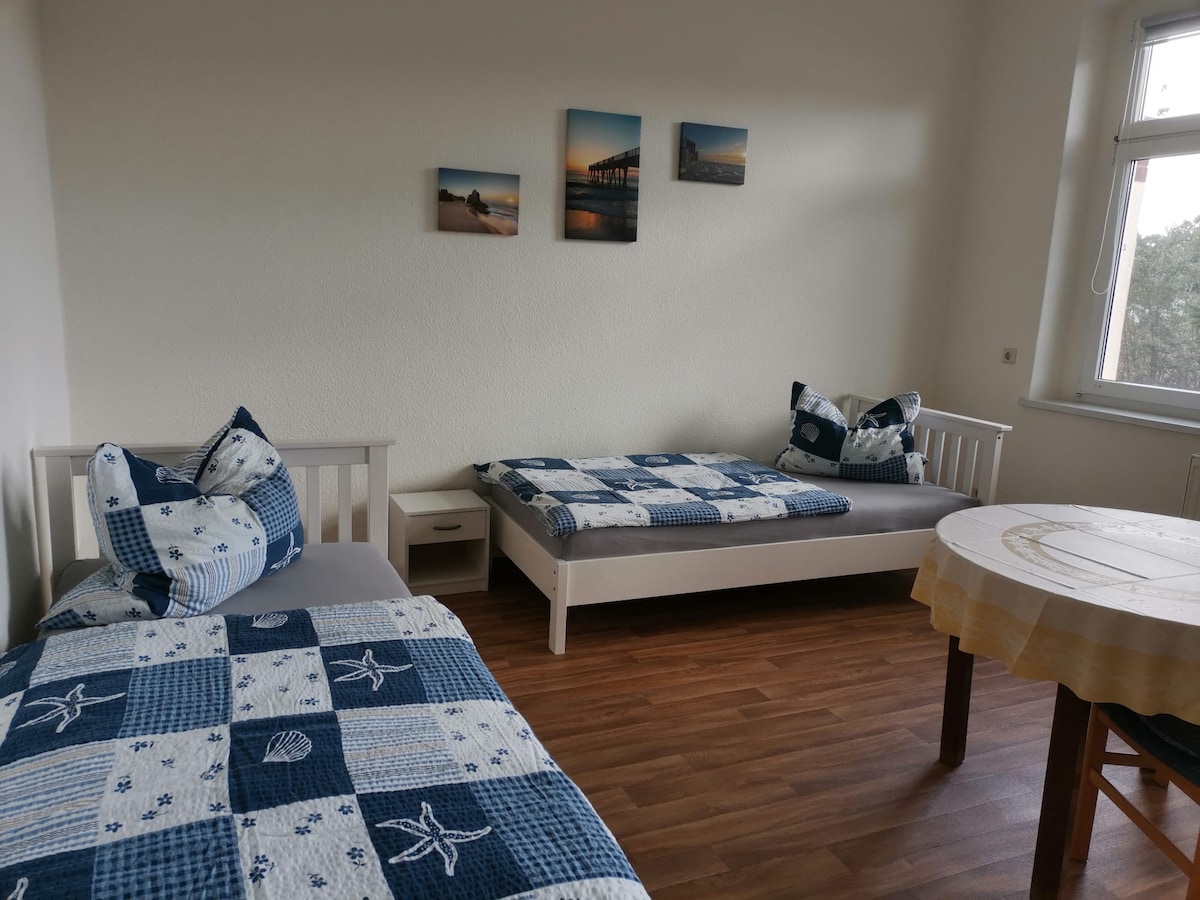 The room contains two single beds covered with patterned blue and white bedding. A small nightstand is positioned between the beds. A dining table with a decorative tablecloth sits near a window, allowing natural light to illuminate the space. Three framed images are displayed on the wall.