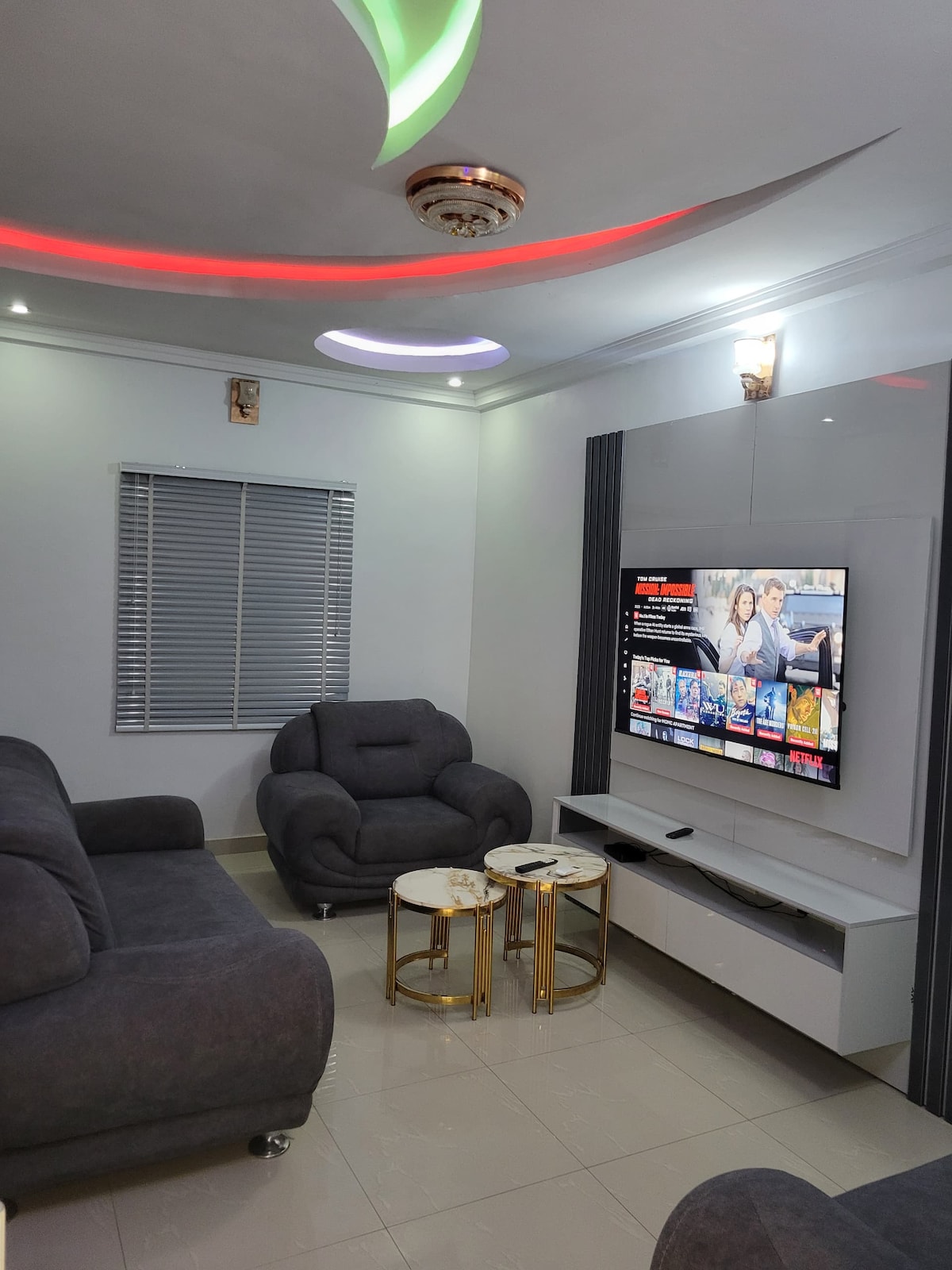 A modern living room is featured, furnished with a grey sofa set and two circular side tables. A large flat-screen television is mounted on the wall, displaying vibrant on-screen content. Soft lighting is provided by recessed lights and a decorative ceiling fixture.