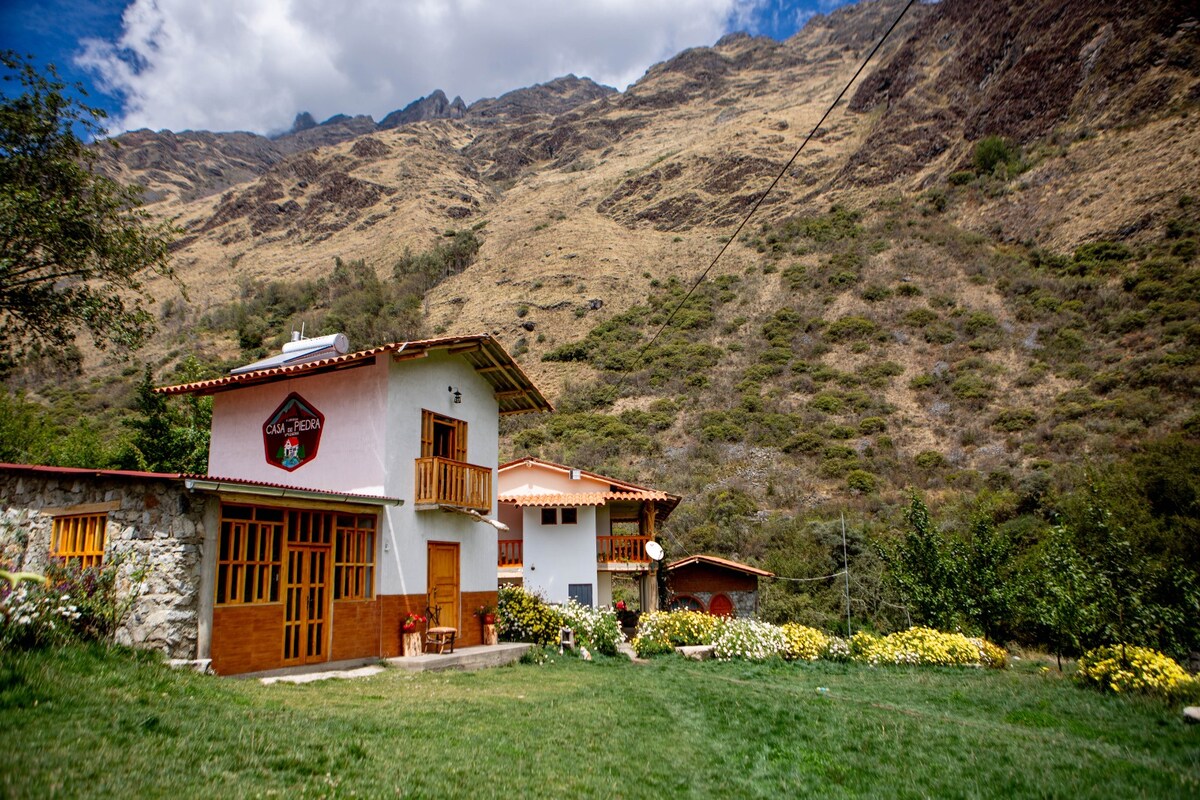 A two-story building features a combination of white walls and stone accents, surrounded by lush greenery and blooming flowers. The backdrop consists of rolling hills and a mountainous landscape, under a partly cloudy sky.