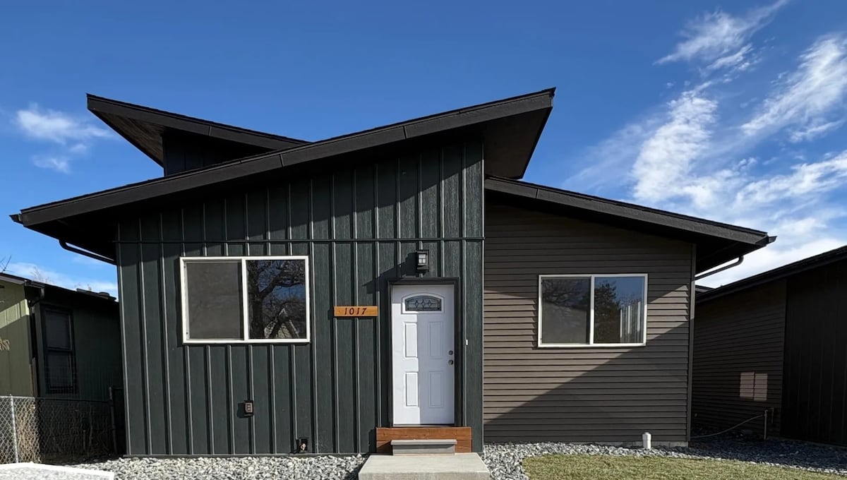 The exterior of the home features a modern design with contrasting dark green and gray siding. A central white door is framed by two large windows. The sloped roof enhances the architectural interest. A gravel path leads to the entrance, and a clear blue sky is visible overhead.