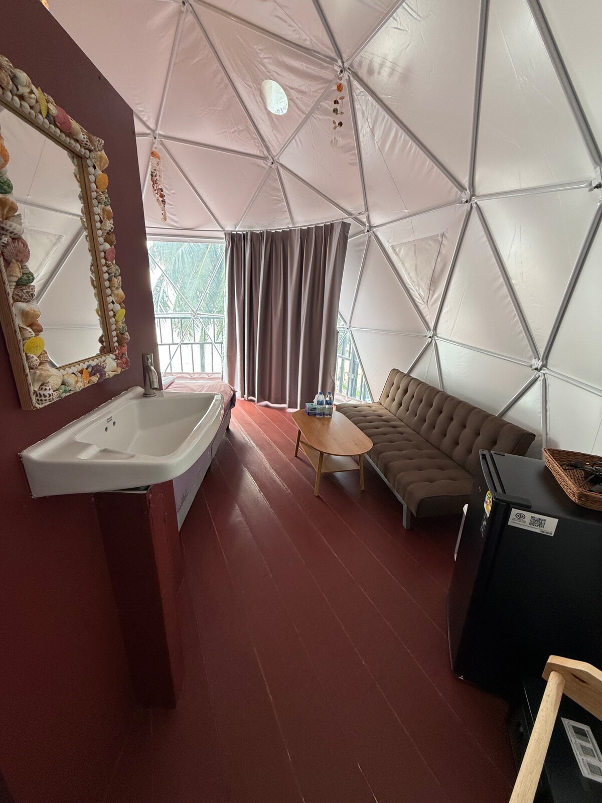 An open interior of a geodesic dome features soft lighting and a spacious seating area with a comfortable couch. A wooden coffee table sits nearby, complemented by a small fridge and a decorative mirror. Floor-to-ceiling curtains frame a bright window, offering a view of the outdoors.