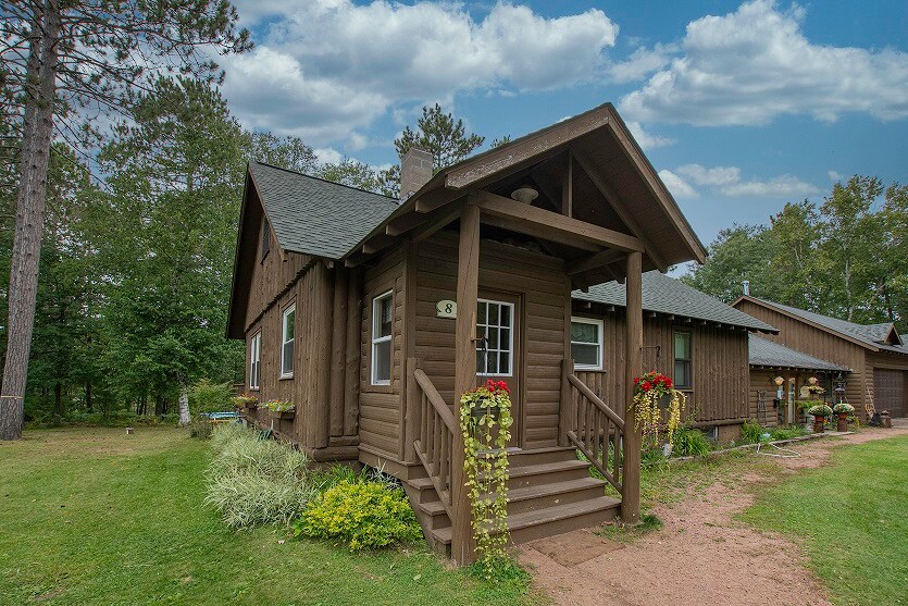 A charming log cabin is shown, nestled among lush greenery. The front porch is adorned with hanging flower baskets, and a pathway leads up to the door. The structure features a sloped roof and multiple windows, providing a rustic yet inviting appearance.