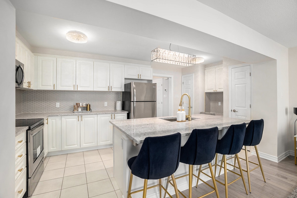 The kitchen features modern white cabinetry and granite countertops, complemented by a stylish light fixture. A kitchen island with seating for four is highlighted, providing a functional space for meal preparation. Stainless steel appliances are visible, enhancing the contemporary design.