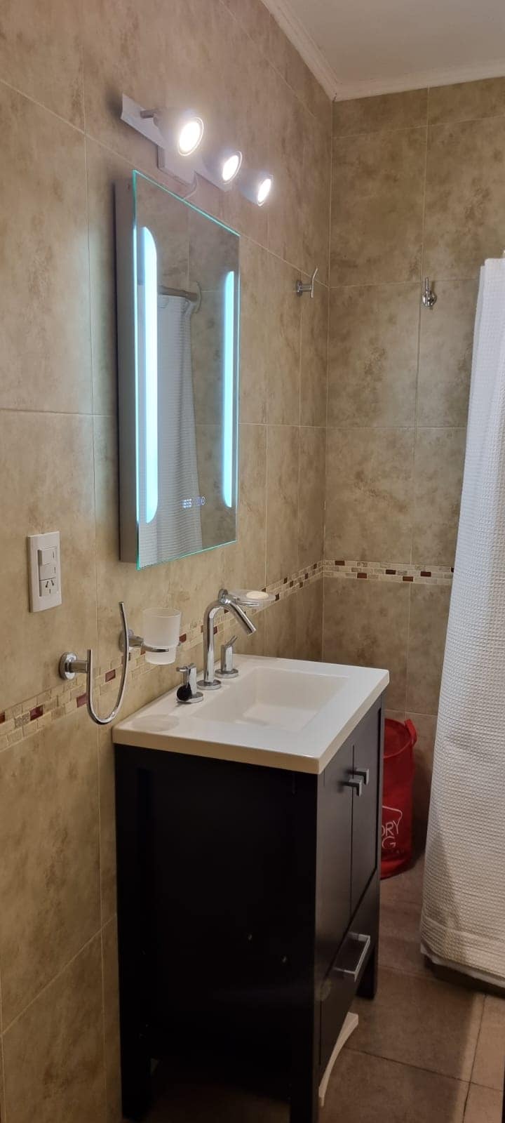 A clean and modern bathroom features a compact vanity with a sink and a large mirror above it. Soft lighting illuminates the space. A shower is enclosed with a white curtain, and tiled walls offer a neutral backdrop.