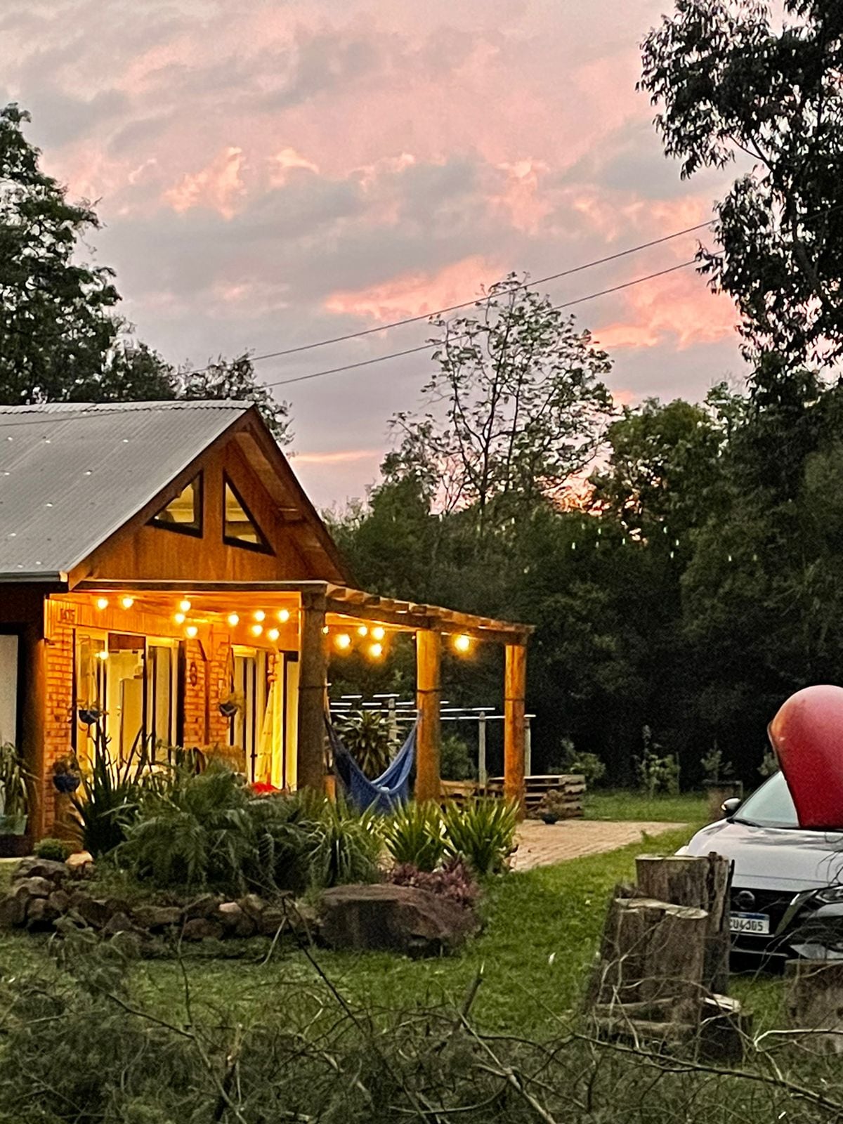 A charming cabin is illuminated by warm string lights, creating a welcoming atmosphere during twilight. The front porch, adorned with a hammock, is surrounded by greenery and features a pathway leading to an outdoor space. Faint hints of a sunset add color to the sky.