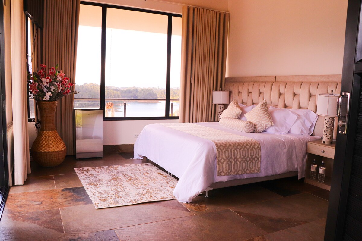 A spacious bedroom is presented with a king-size bed adorned in light bedding. Large glass windows offer panoramic lake views, while soft curtains frame the scene. A decorative vase with flowers is placed beside the bed, and a subtle area rug is positioned on the stone flooring.