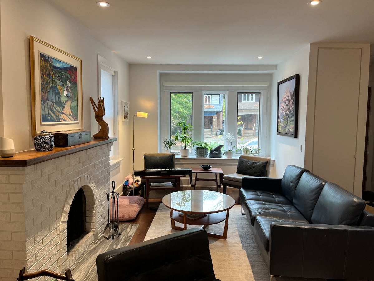 A modern living room is showcased, featuring a round coffee table centered on a light rug. A comfortable leather sofa and chairs are positioned around the table. Natural light floods through large windows, illuminating the artwork on the walls and the minimalist decor.