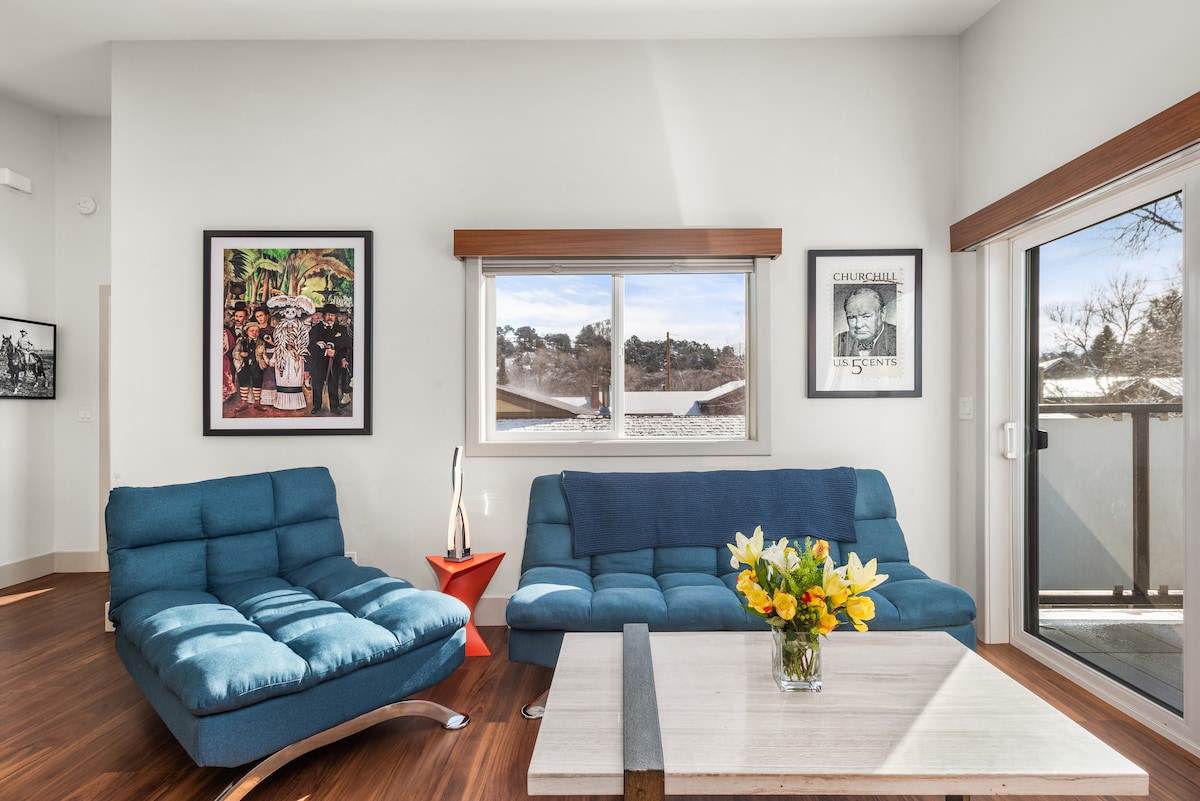 A bright and inviting living space features two modern, blue lounge chairs and a low coffee table adorned with a bouquet of yellow flowers. Large windows allow natural light to fill the room, complemented by framed artwork on the walls. A sliding glass door provides access to an outdoor area.