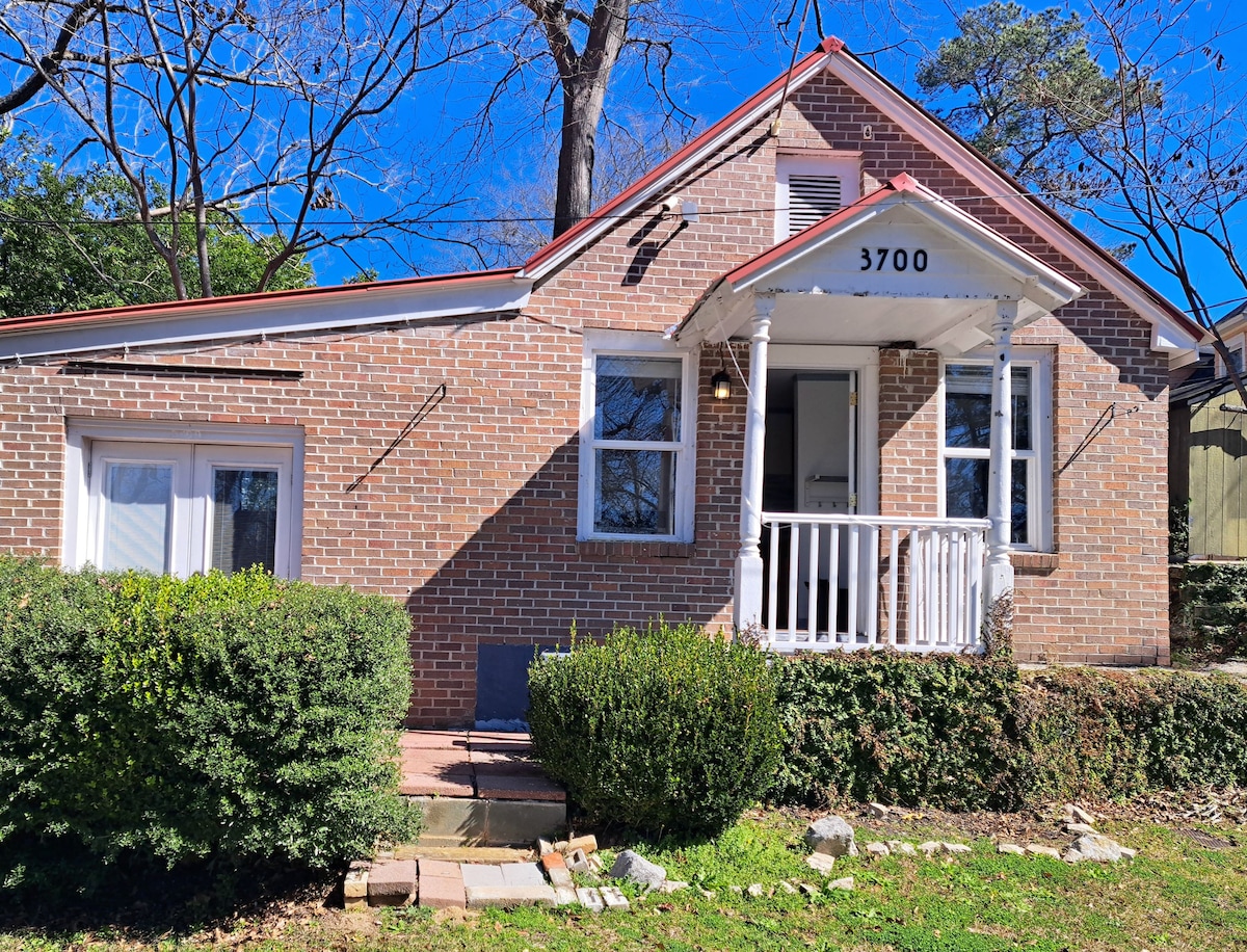 The exterior of a charming brick house is showcased, featuring a welcoming entrance with a covered porch. A clearly marked house number is displayed above the door. Surrounding greenery and low shrubs enhance the outdoor space. The roof is complemented by a bright red metal accent.