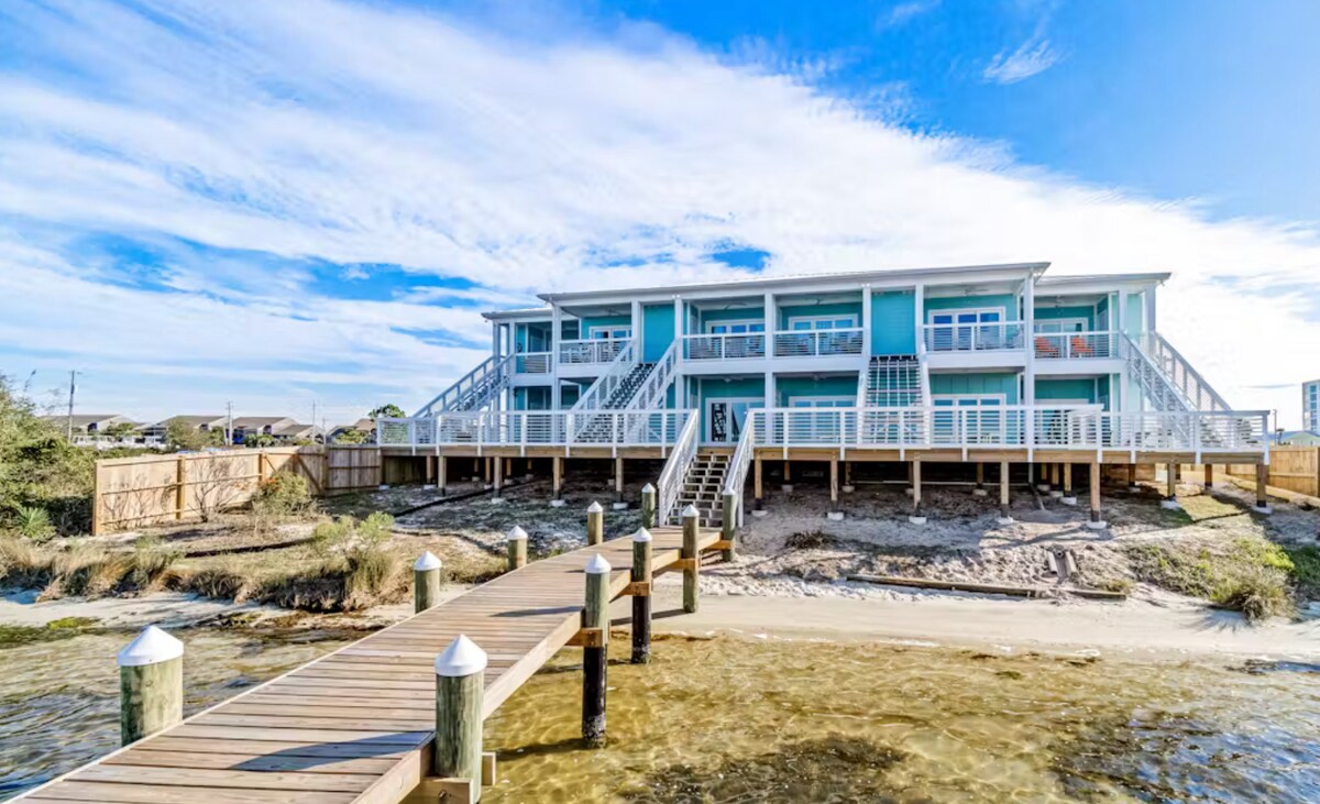 A waterfront condo building with multiple levels is depicted, featuring large windows that reflect sunlight. A wooden dock extends toward the sandy shore, where the gentle waves lap against the pilings. Clear skies and a sense of tranquility are present in the scene.