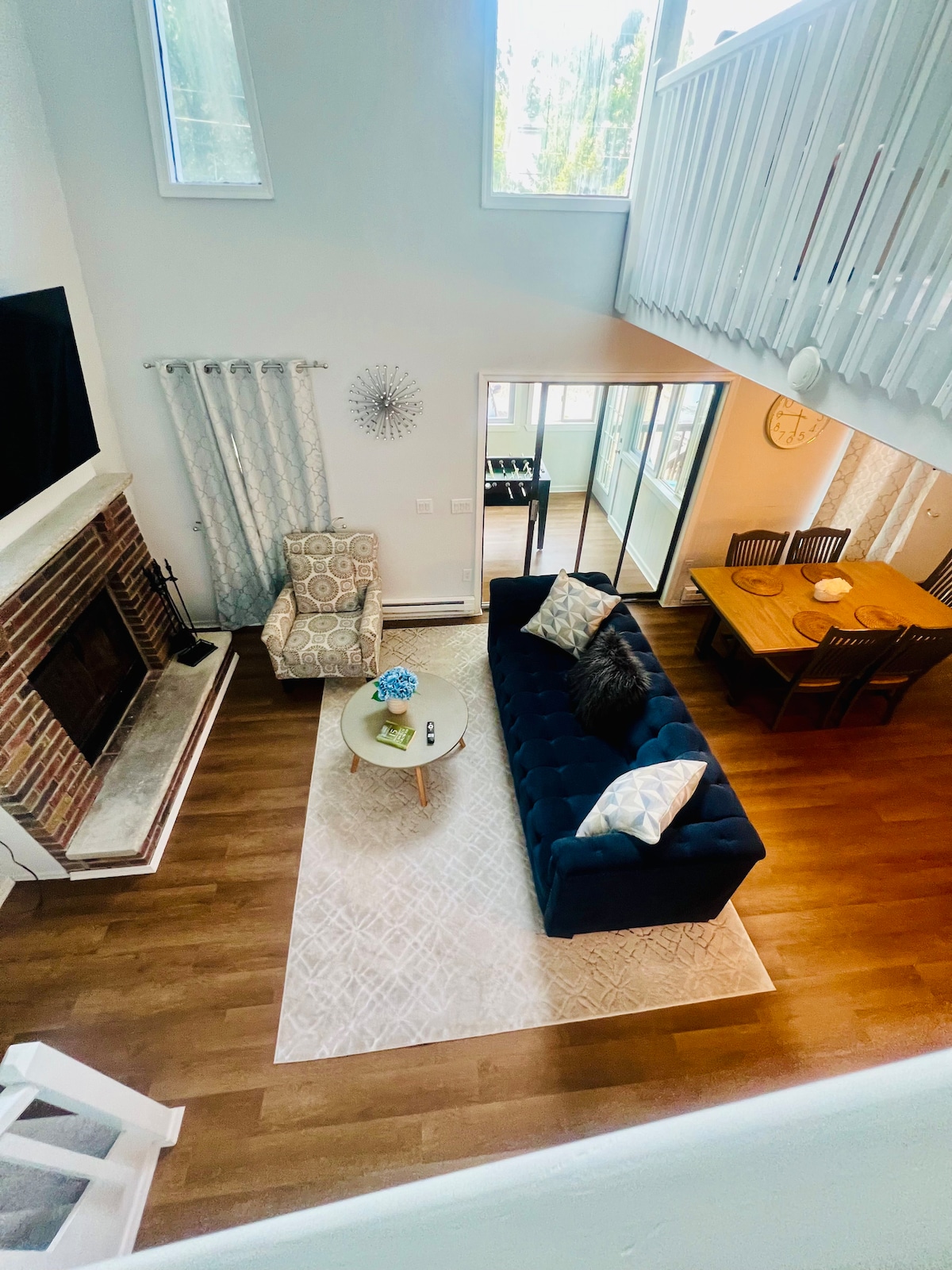 An open living area features a navy blue sofa with decorative pillows, a round coffee table, and a cozy chair. A large fireplace is visible against a brick wall. Natural light floods the space through high windows, illuminating a dining table positioned nearby.