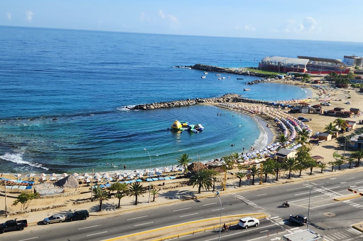 A wide view of a sandy beach along the coastline is visible, with calm blue waters gently lapping at the shore. Colorful beach umbrellas line the shore, while small boats can be seen floating in the distance. The horizon features a clear sky, enhancing the vibrant coastal scene.