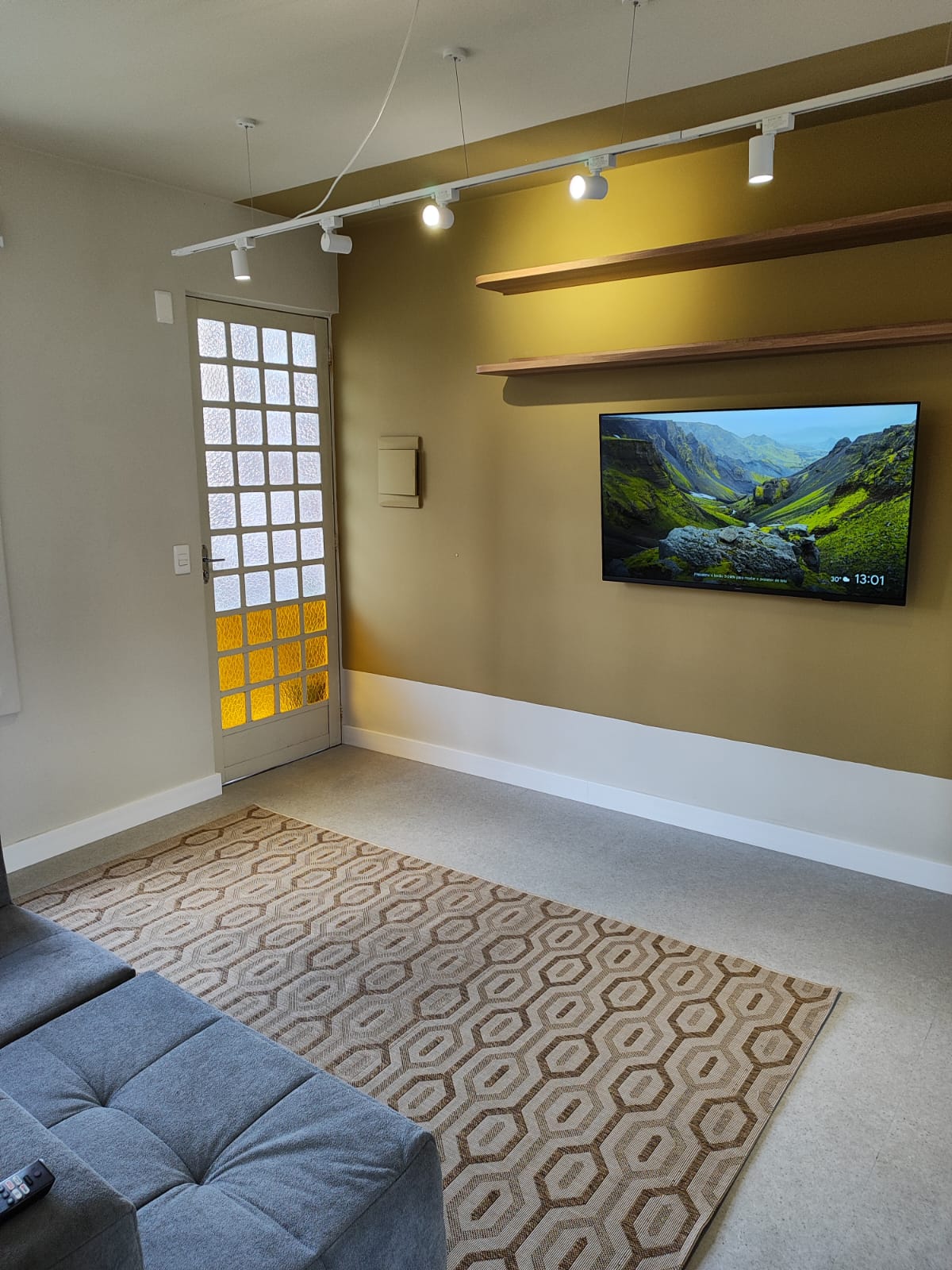 A cozy living area features a large television mounted on a mustard-colored wall, complemented by a warm-toned rug on the floor. Natural light streams in through a glass panel door, enhancing the inviting feel of the space. Simple shelving is present above the television.