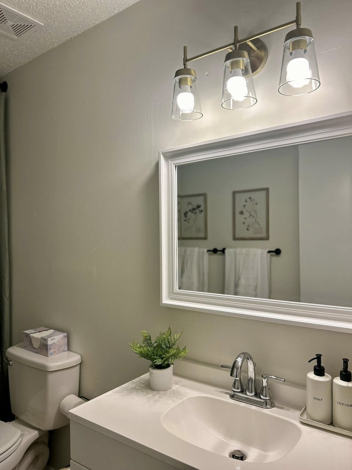 A well-lit bathroom features a white vanity with a sink, complemented by modern fixtures. A large mirror is framed in white, reflecting the space. Two framed artwork pieces are displayed above neatly hung white towels on a black rack. Small potted greenery adds a touch of color.