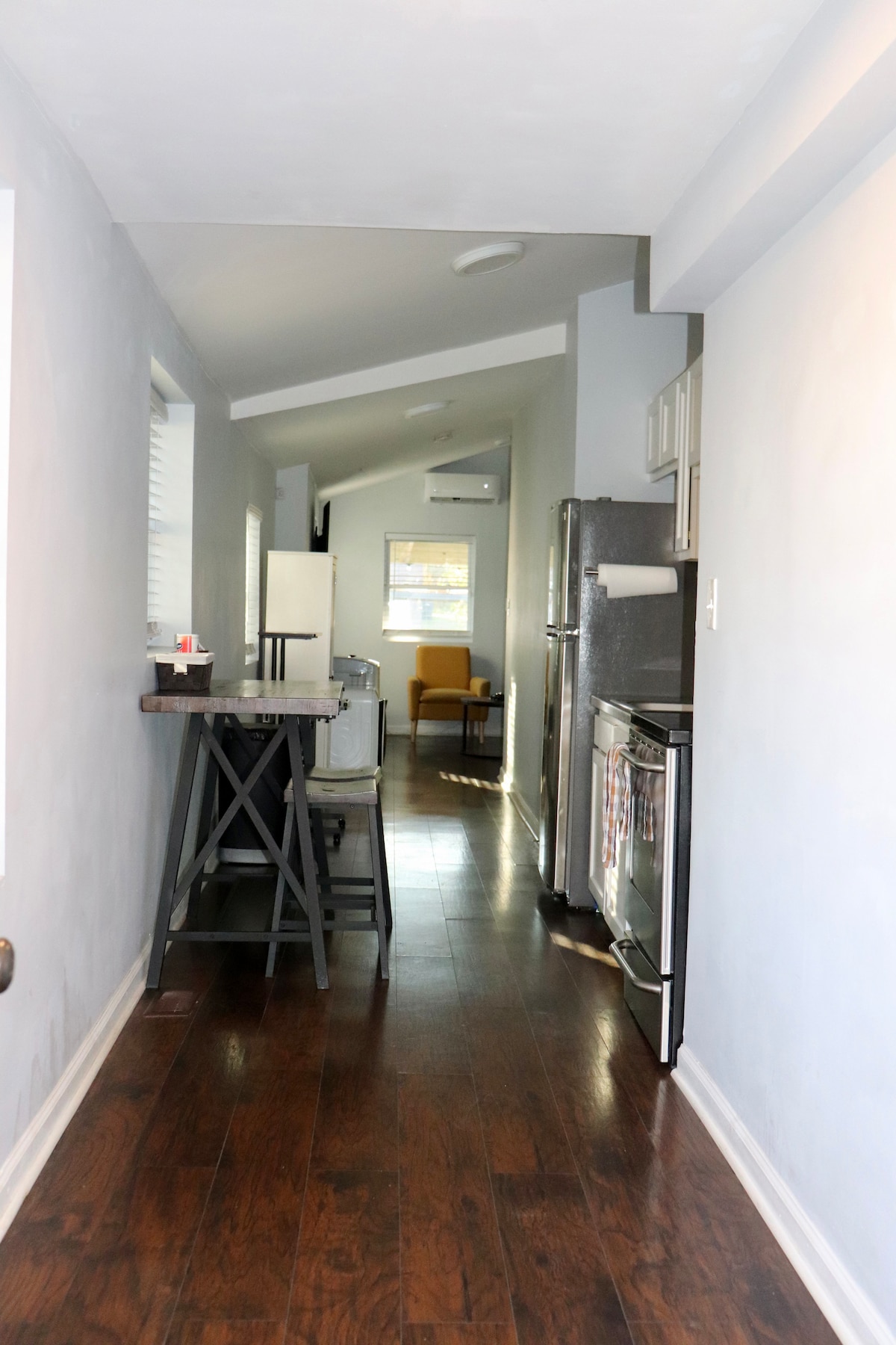 A narrow hallway is shown with light-colored walls and hardwood flooring. A small dining table with two stools is situated to one side, while modern appliances are visible in the kitchen area. A window allows natural light to fill the space, along with a glimpse of seating beyond.
