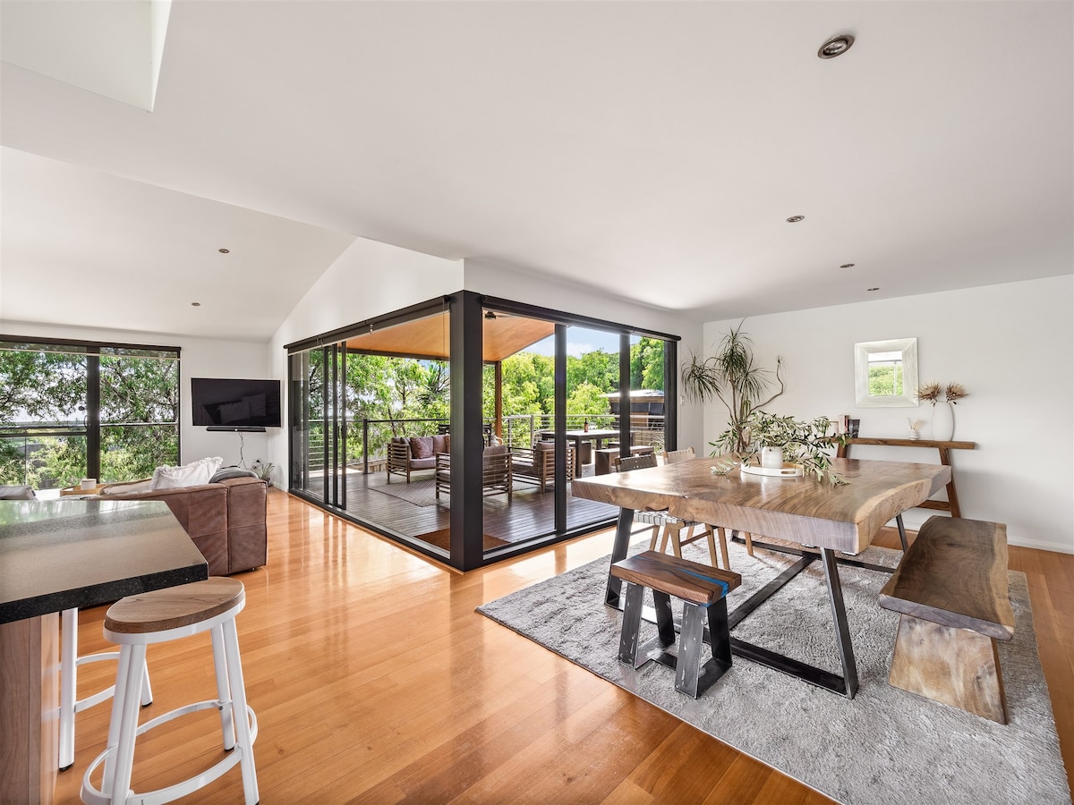 An open-plan living area features a wooden dining table surrounded by benches, set against a backdrop of large glass sliding doors that lead to an outdoor deck. Natural light fills the space, highlighting the polished wooden floors and modern furnishings in a relaxed layout.