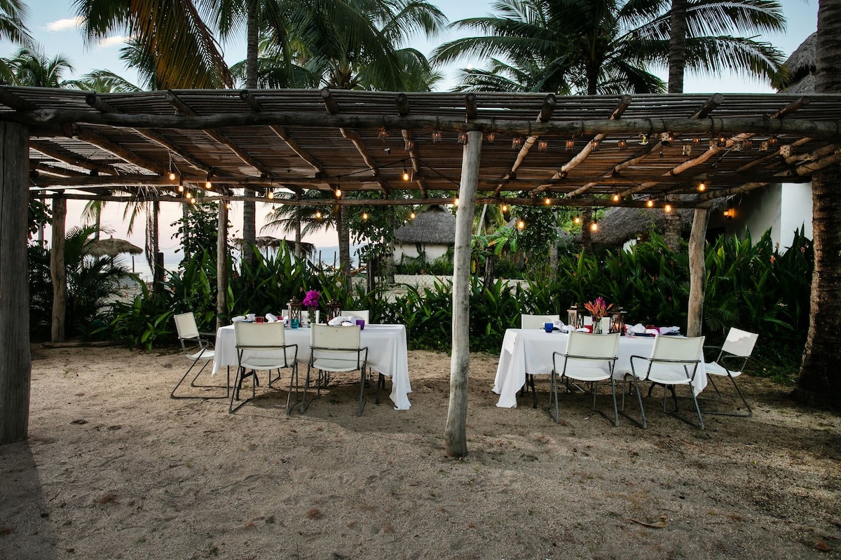 A sandy dining area is set under a wooden pergola, featuring white tablecloths on several tables. Soft lighting from string lights hangs above, surrounded by lush tropical vegetation. The tranquil atmosphere is enhanced by palm trees swaying gently in the background.