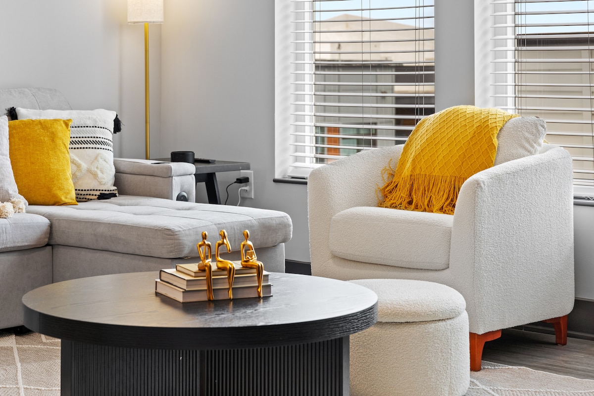 A bright living area features a sectional gray sofa with black and white accent pillows. A round black coffee table is arranged with stacked books and decorative figurines. A textured white armchair with a yellow throw sits nearby, complemented by natural light from window coverings.
