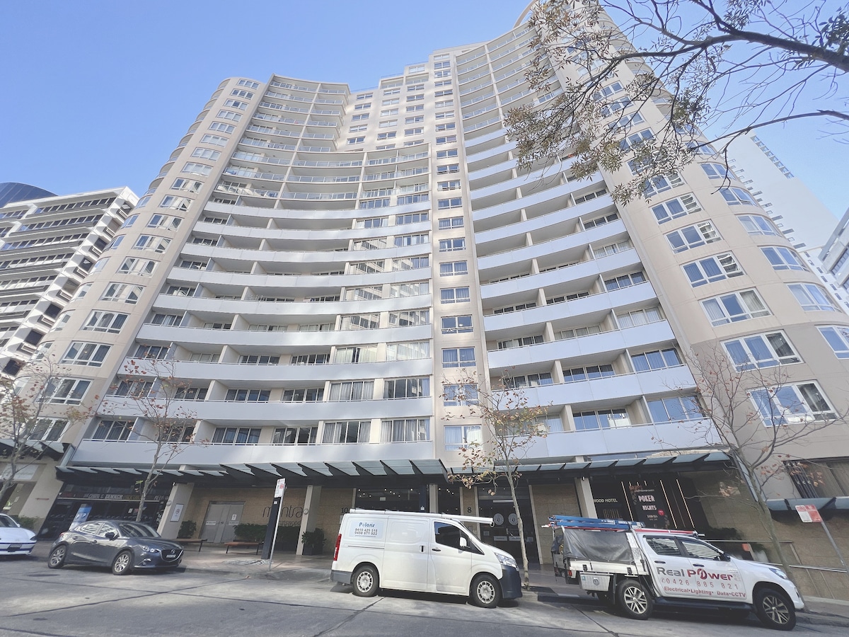 The exterior of the Mantra Chatswood building is displayed, featuring a modern design with curved balconies and large glass windows. The entrance is framed by awnings, and there are vehicles parked along the street in front.